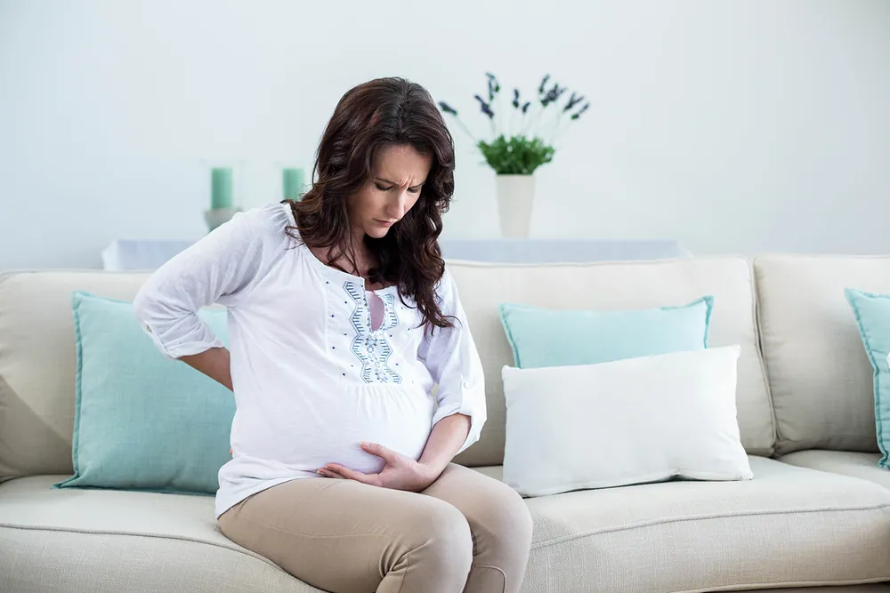 woman having a lower back pain while being pregnant and sitting in the sofa
